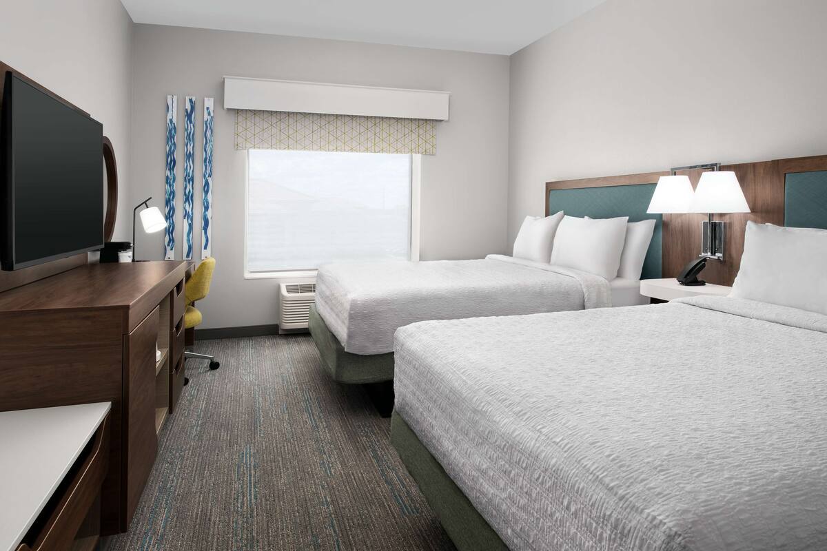 A hotel room with two beds, a desk, a TV, and a window with curtains.