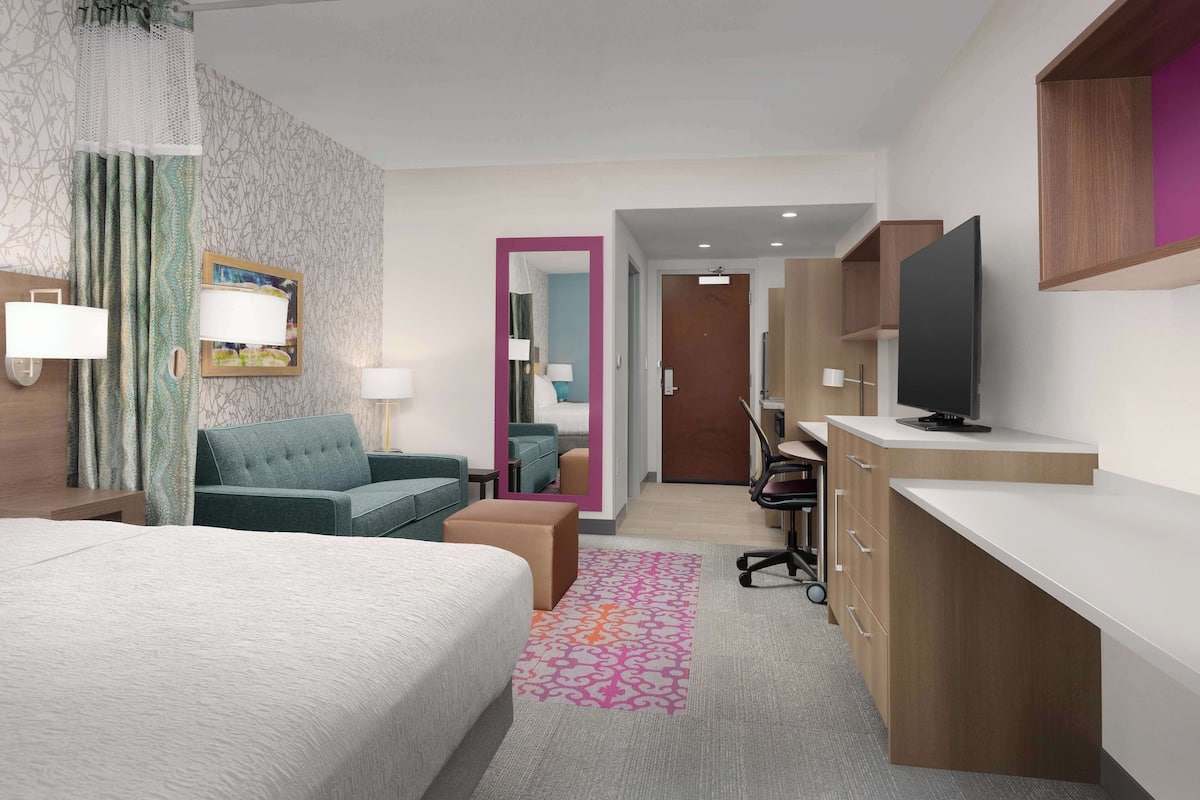 Habitación de hotel con cama, sofá, escritorio, televisión y baño visible a través de una puerta abierta.