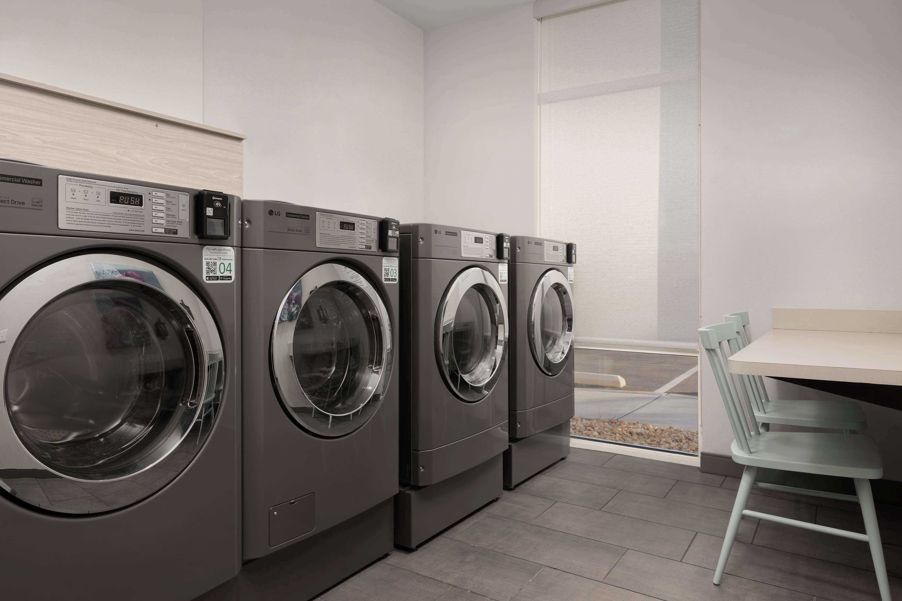 A row of commercial washers in a clean, modern laundry room with a small table and chair.