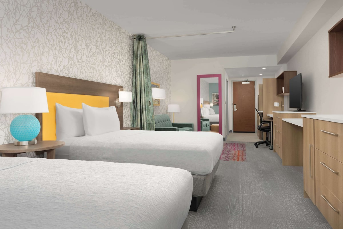 Habitación de hotel con dos camas, televisión, escritorio y baño visible a través de una puerta abierta.