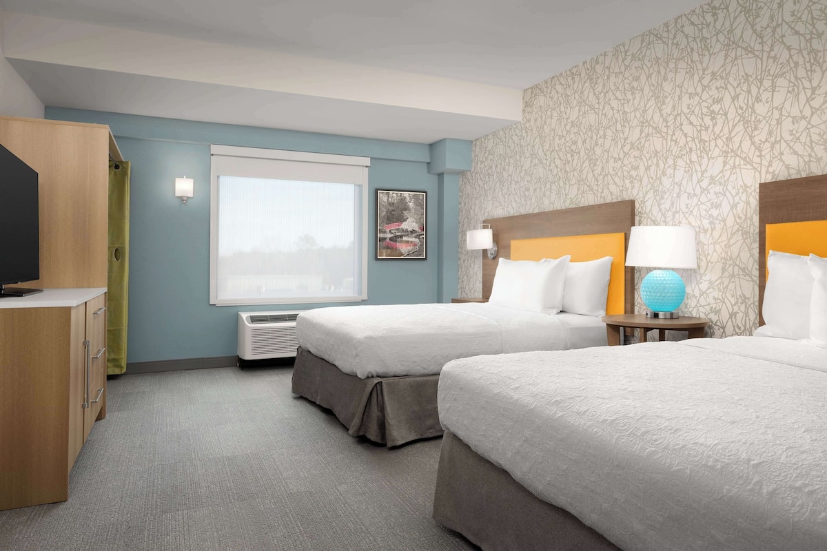 Habitación de hotel con dos camas, televisor, escritorio y ventana con persianas.