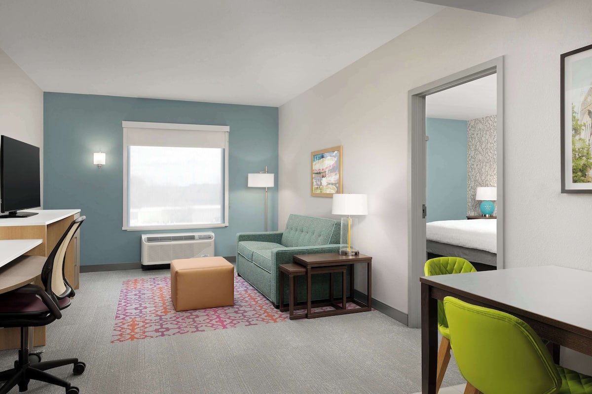 Una habitación de hotel compacta con escritorio, sofá verde, mesita, lámpara y cama.