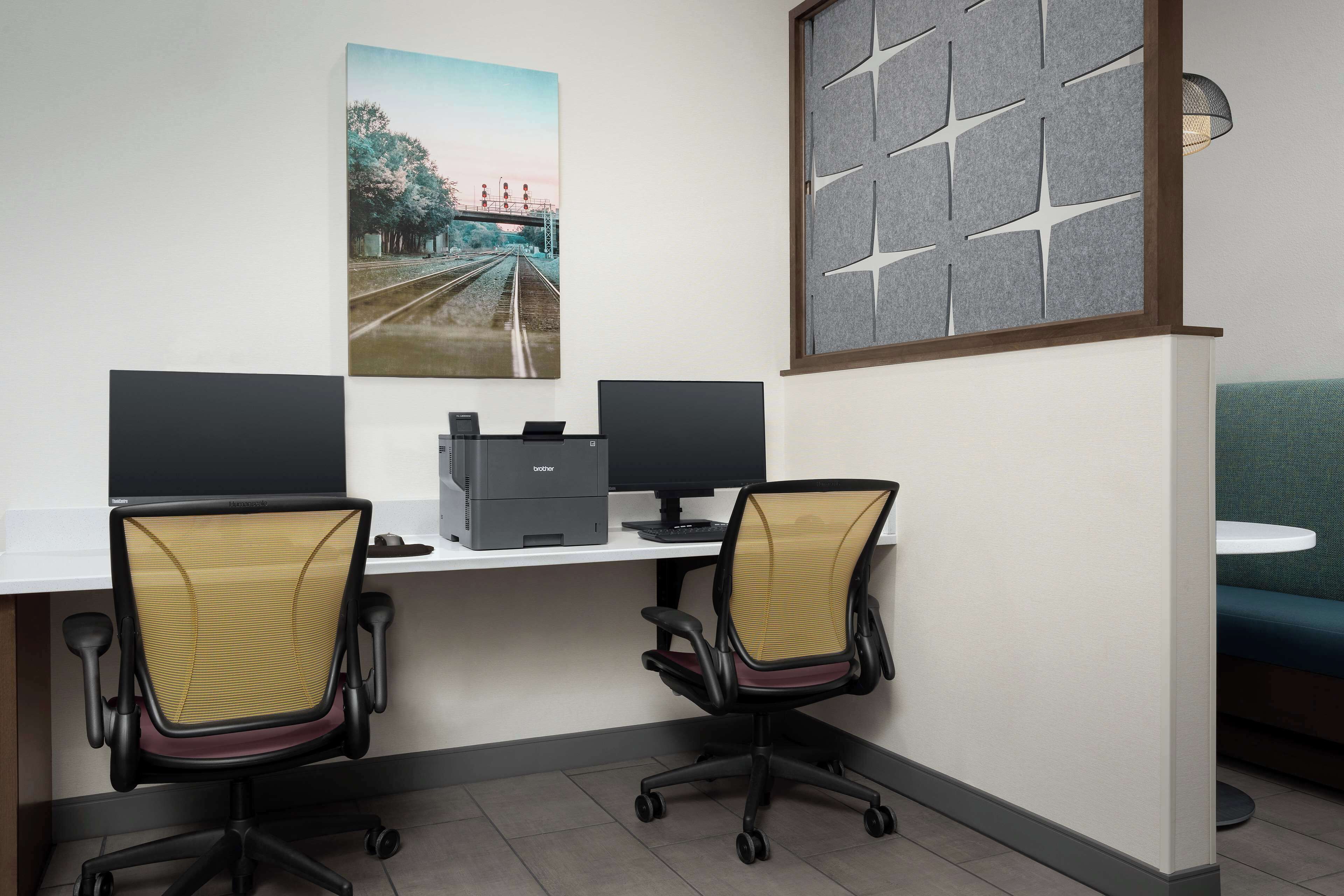 A modern office space with two desks, each with a computer monitor, a printer, and an ergonomic office chair. There is a large window with a view of a bridge and trees.
