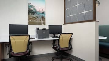 A modern office space with two desks, each with a computer monitor, a printer, and an ergonomic office chair. There is a large window with a view of a bridge and trees.