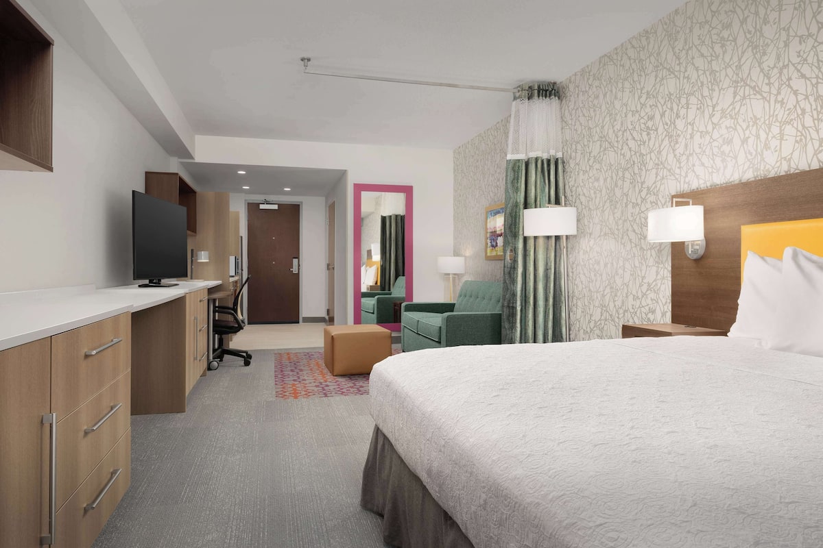 Habitación de hotel con una cama grande, un escritorio con computadora, una silla, un televisor y una puerta que da a otra habitación.