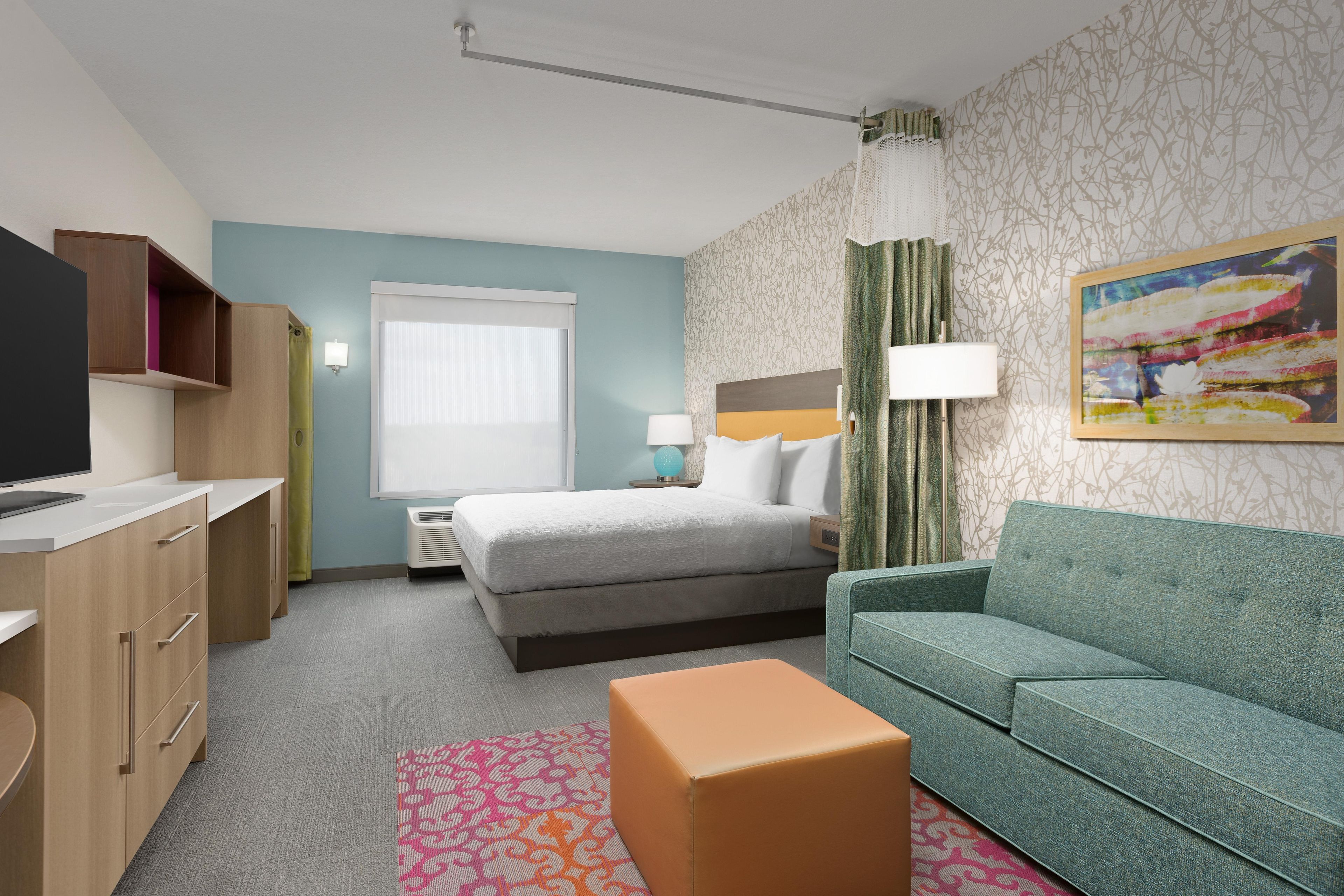 A hotel room with a bed, a desk with a flat-screen TV, a green sofa, and a patterned rug.