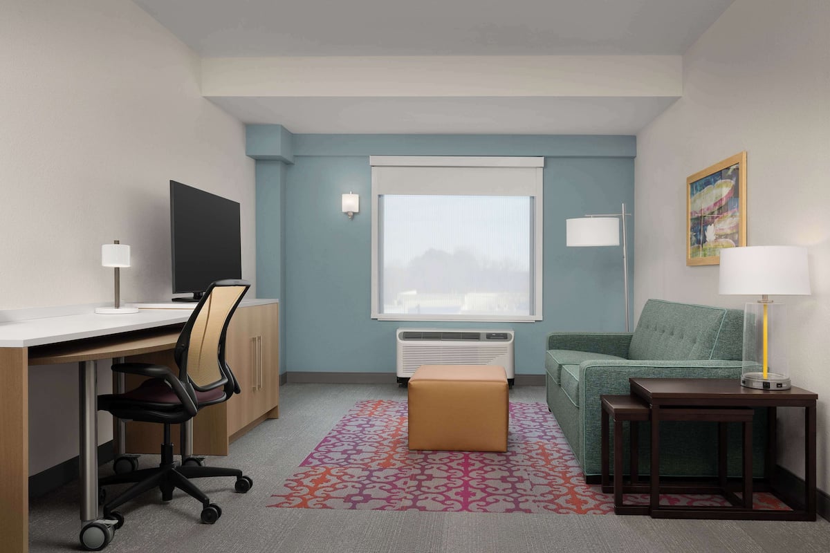 Habitación de hotel con escritorio, silla de oficina, sofá, reposapiés, mesita, lámpara, aire acondicionado y ventana con persianas.