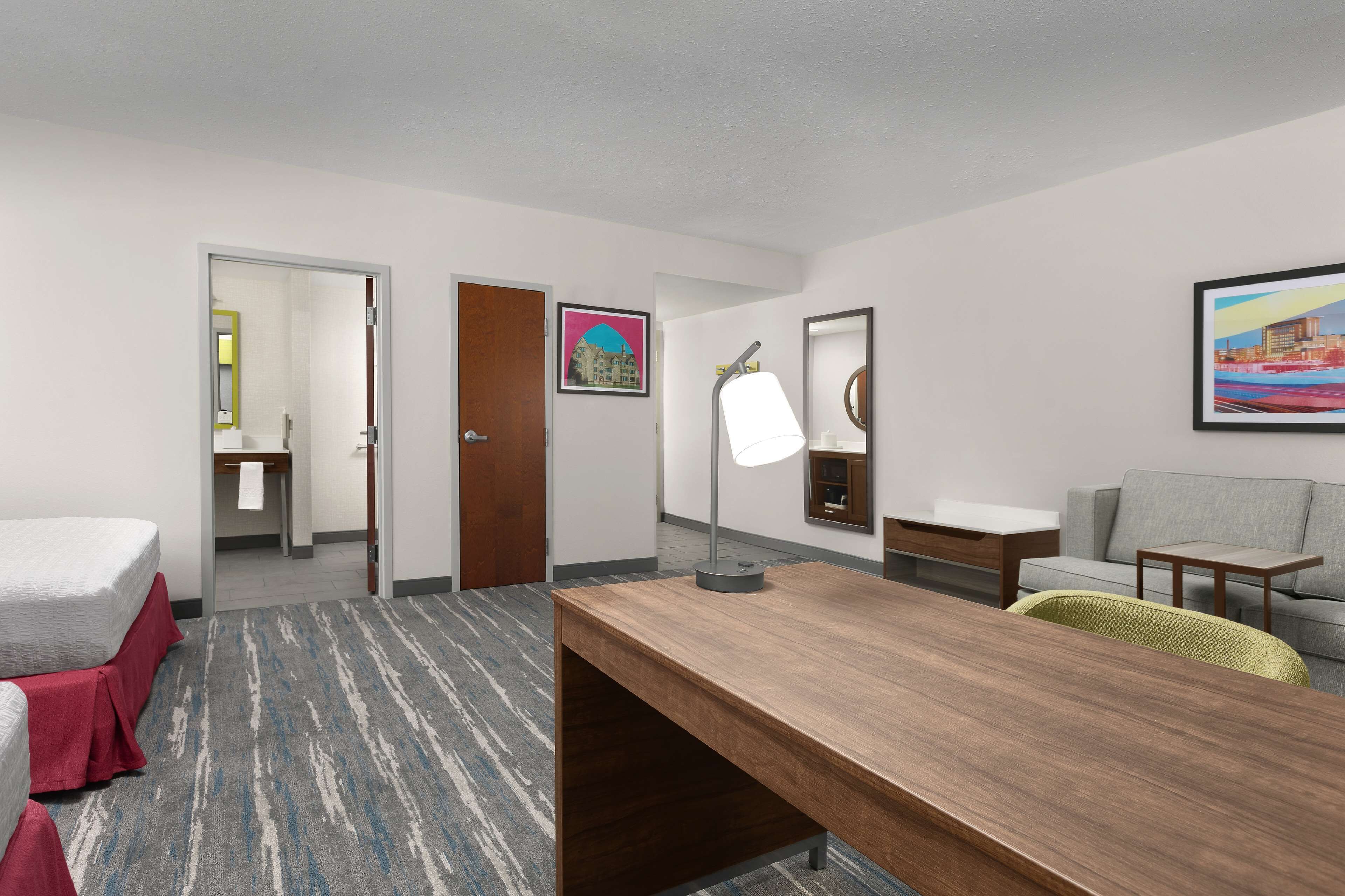 A hotel room with a bed, a desk, a chair, a sofa, a lamp, and a painting on the wall.