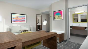 Habitación de hotel con escritorio, sofá, cama, baño con espejo y lavamanos, y dos cuadros en las paredes.