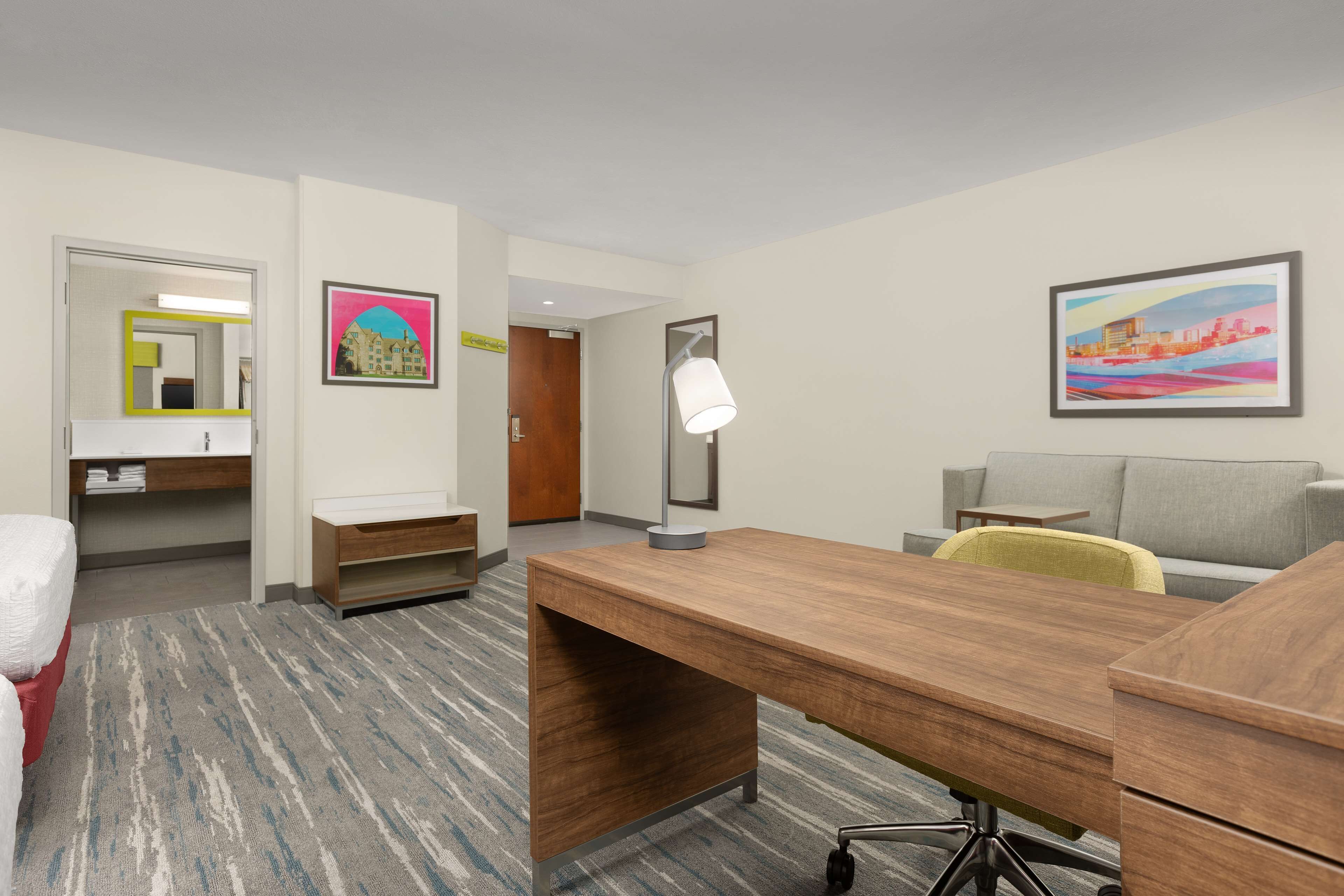 A hotel room with a wooden desk, a chair, a bed, a bathroom with a mirror, and two paintings on the walls.