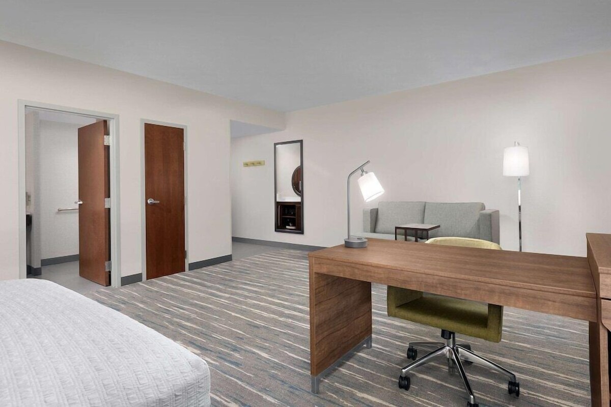 A hotel room with a wooden desk, a grey sofa, a lamp, and a patterned carpet.