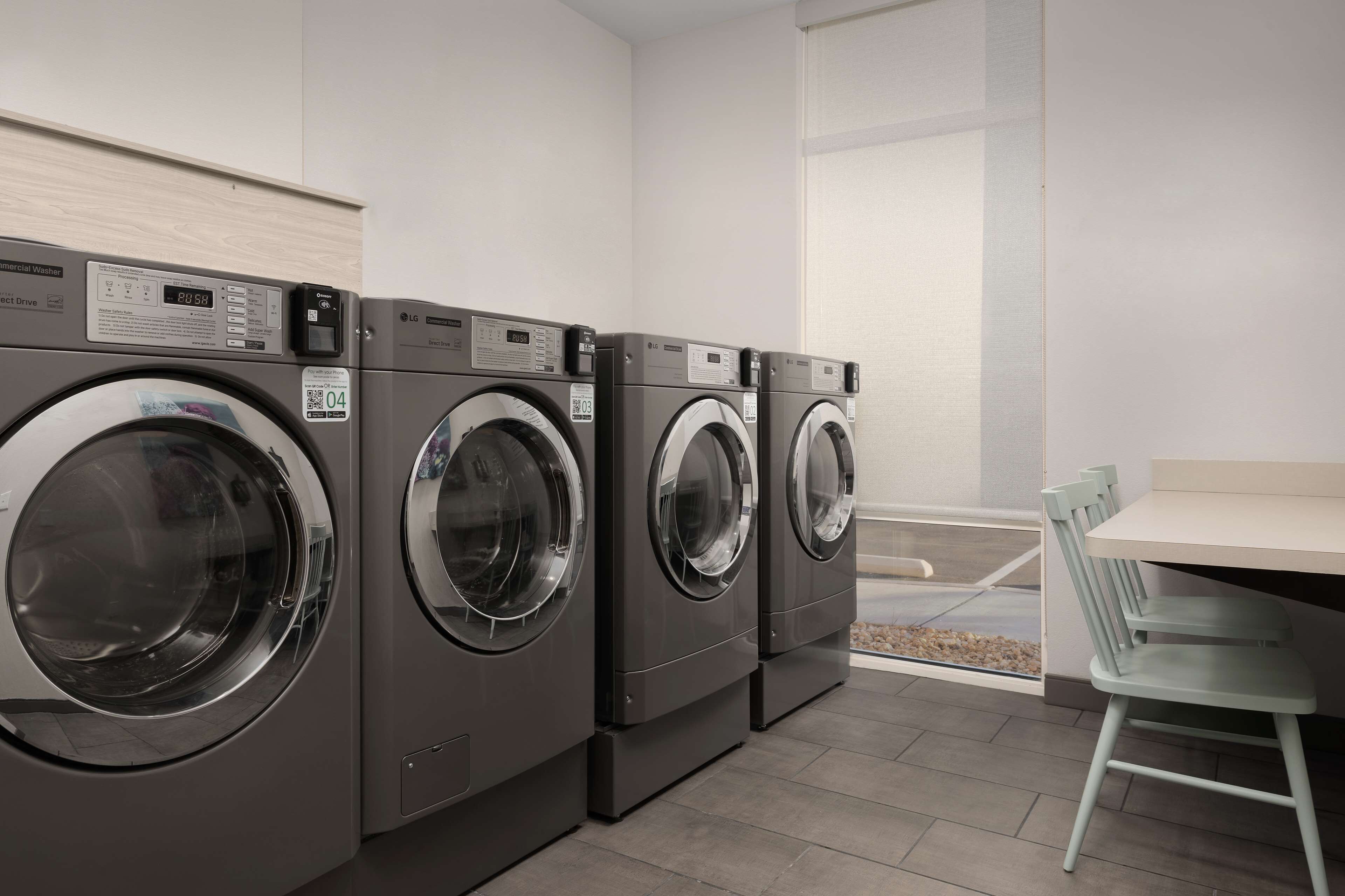 A row of commercial washers in a clean, modern laundry room with a small table and chair.