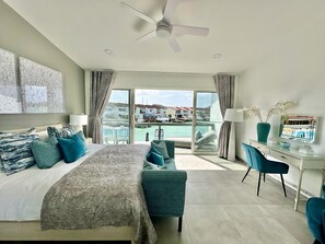 A hotel room with a bed, a desk, a chair, a balcony with a view of houses and water, and a ceiling fan.