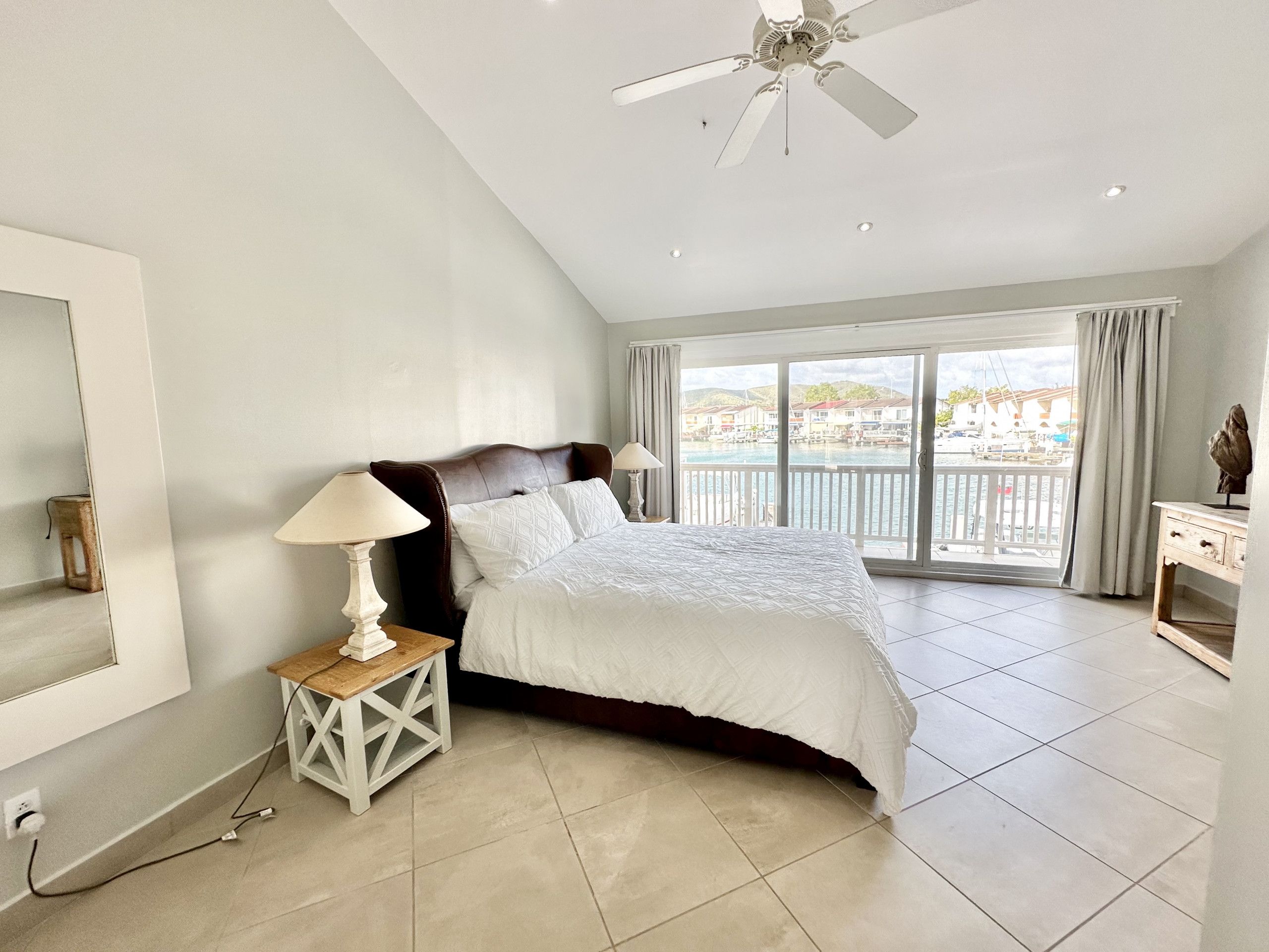 A bedroom with a large bed, a ceiling fan, a nightstand with a lamp, and a view of a body of water through the window.