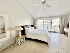 A bedroom with a large bed, a ceiling fan, a nightstand with a lamp, and a view of a body of water through the window. - Enchanting Waterfront Villa (Jolly Harbour)