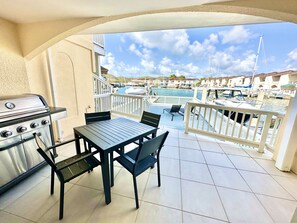 A balcony with a grill, a table, and chairs, overlooking a marina with boats and buildings. - Enchanting Waterfront Villa (Jolly Harbour)