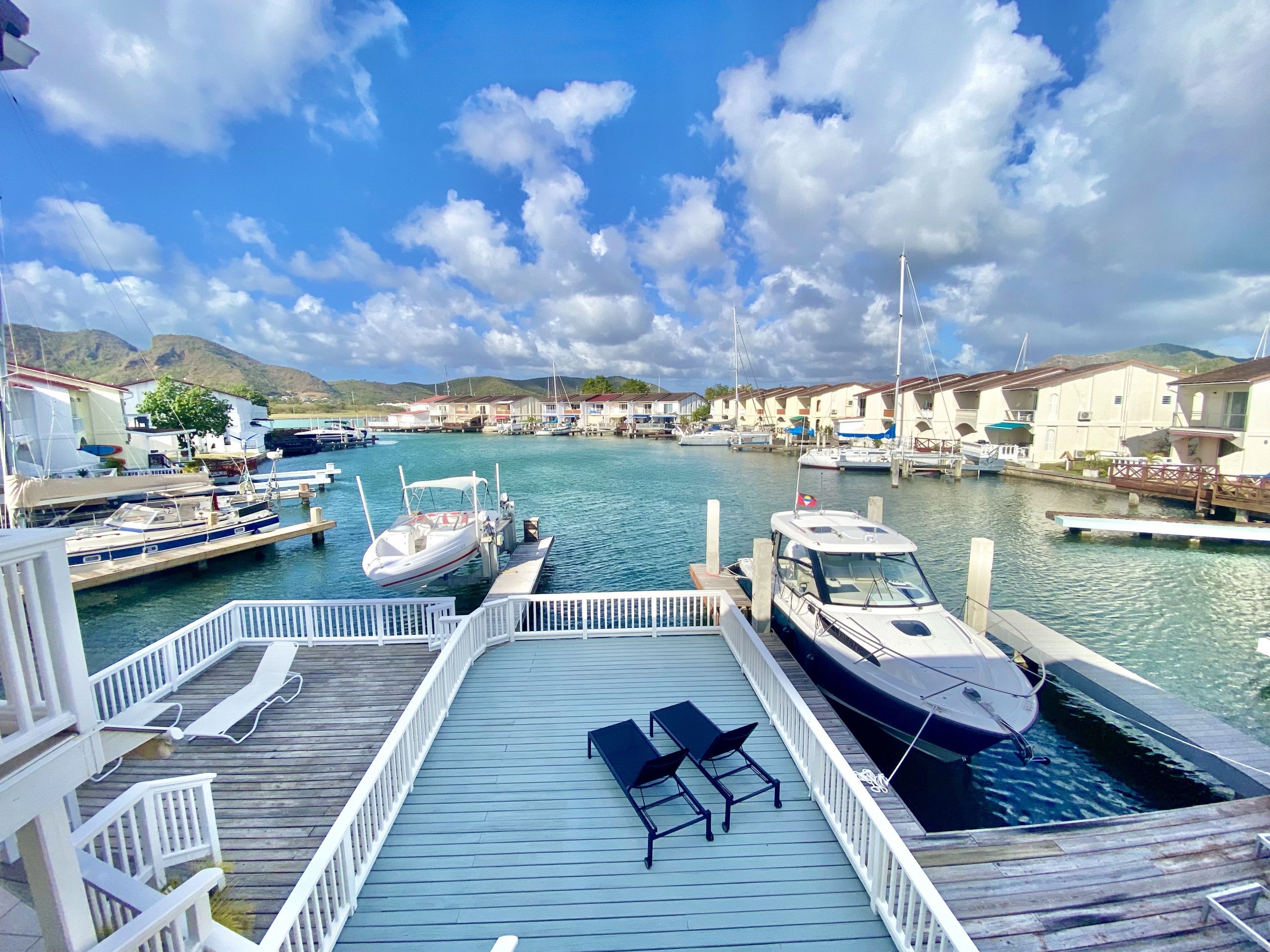 A marina with boats docked, a wooden deck with lounge chairs, and buildings in the background.