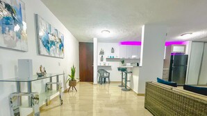 Una sala moderna con una mesa de centro de vidrio, una planta en maceta y un comedor con taburetes de bar.