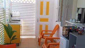 Una cocina compacta con armarios blancos y una pequeña encimera. Hay dos sillas anaranjadas y una silla amarilla cerca de la cocina. Una mesa con mantel colorido está en primer plano.