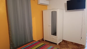 Una habitación con un armario blanco, una cortina gris, un tapete de rayas coloridas y un televisor de pantalla plana montado en la pared.