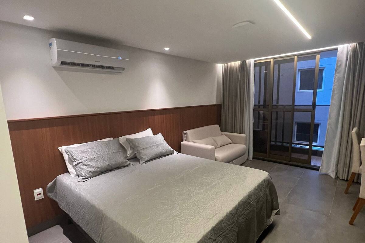 A modern hotel room with a large bed, a sofa, a sliding glass door leading to a balcony, and an air conditioning unit on the wall.