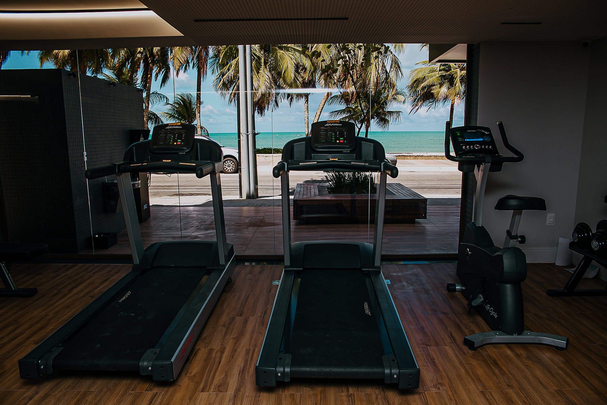 Ein Fitnessraum mit Laufbändern und einem Ergometer, große Fenster mit Blick auf den Strand und Palmen.
