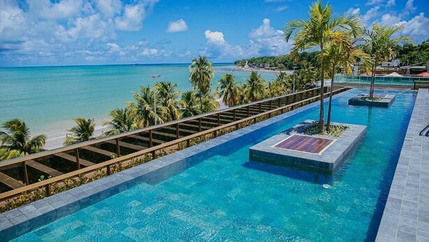 A pool with a wooden deck and palm trees, overlooking a beach and ocean. - Unity Suites (João Pessoa)