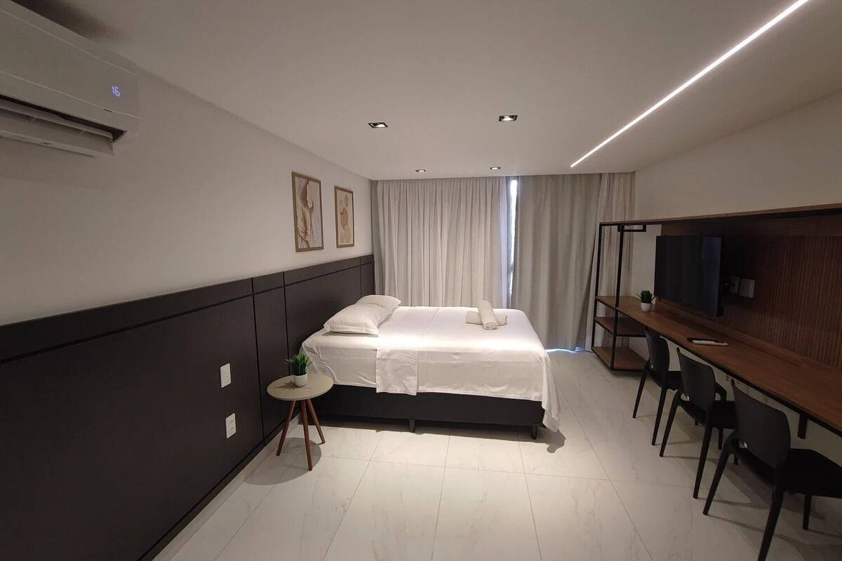 A modern hotel room with a bed, a desk, a chair, a TV, and a small plant.