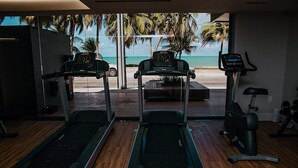 A gym with treadmills and an exercise bike, large windows with a view of the beach and palm trees. - Unity Suites (João Pessoa)