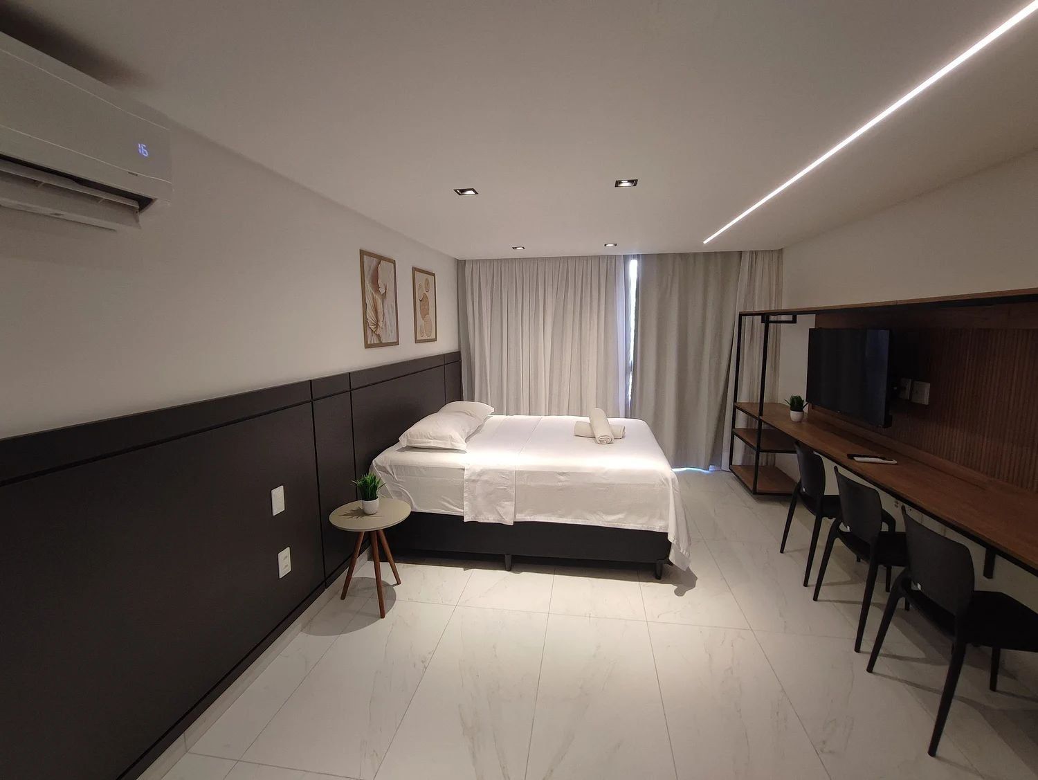 Quarto de hotel moderno com cama, escrivaninha, cadeira, televisão e uma pequena planta.