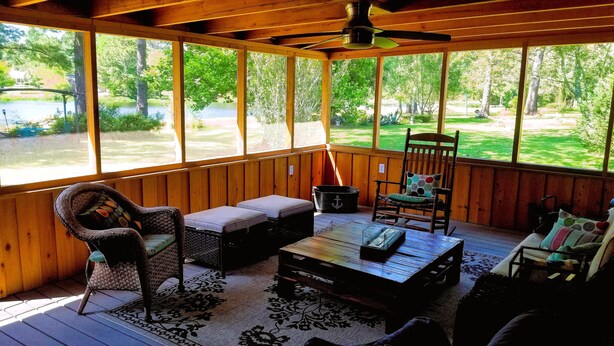 A covered porch with wooden walls, a ceiling fan, wicker furniture, and a view of a lake and greenery.