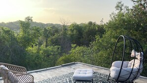 Un área de estar al aire libre con mobiliario de mimbre, una silla colgante y un tapete estampado en una terraza con vistas a árboles.