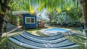 En hängmatta under ett palmträd, en blå byggnad och en swimmingpool.