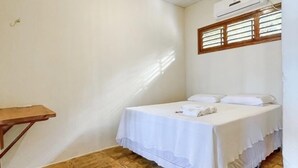 A small, clean room with a bed, a wooden table, and a window with blinds. - Pousada Estrela Peroba (Icapuí)