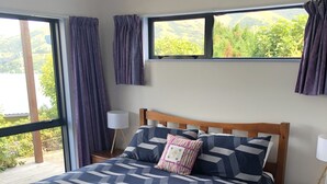 A bedroom with a large bed, a wooden headboard, a window with curtains, a bedside table, and a view of greenery outside. - Peaceful piece of Paradise (Wainui)