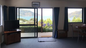 A room with a large sliding glass door leading to a balcony with a scenic view of a lake and mountains. There is a television on a wooden stand and a dining table with chairs. - Peaceful piece of Paradise (Wainui)