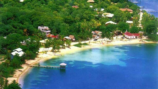 Une zone côtière avec une plage, une eau d’un bleu limpide et plusieurs bâtiments entourés de verdure.