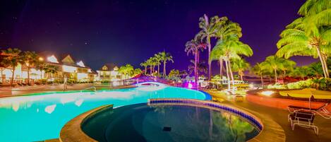 Un área de piscina nocturna con un jacuzzi, palmeras y edificios de hotel al fondo.