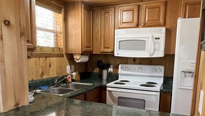 A kitchen with wooden cabinets, a white microwave, a stove, and a refrigerator. There are two stools in the foreground.