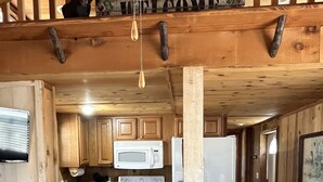A rustic cabin interior with a wooden interior, a kitchen area with a microwave and stove, two wooden stools, and a 'WELCOME' sign above the door.