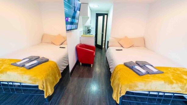 A small, brightly lit room with two beds, each with a yellow blanket and a remote control. A red chair is visible in the center, and a small table with a lamp is against the wall. - Cosy Studio Close to the University (Birmingham)