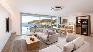 Ein modernes Wohnzimmer mit einer großen Couch, einem Couchtisch und einem Essbereich mit Stühlen und einem Tisch. Es gibt eine Küchenzeile mit einem Kühlschrank und Blick auf das Meer.