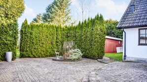 A paved driveway leading to a white building with a dark roof, surrounded by neatly trimmed hedges and a red barn in the background.