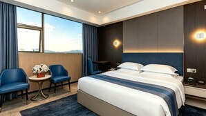 A hotel room with a large bed, a desk, two chairs, a small table with a vase, and a view of the outdoors. - Green Island Holiday Hotel (Jiangmen)