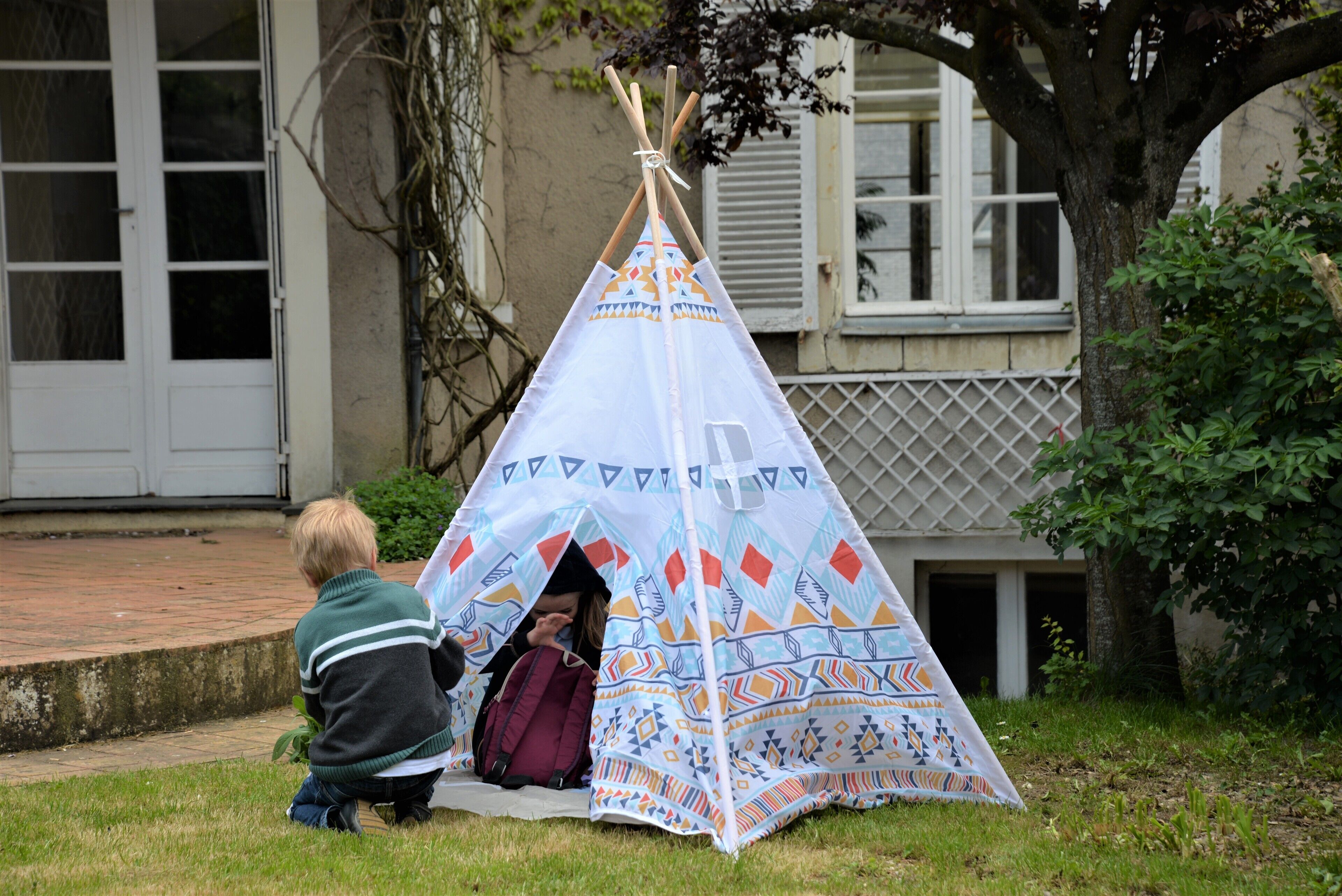 A tent with a geometric pattern, set on grass near a building with a window and a door.