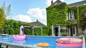 Una piscina al aire libre con juguetes inflables, rodeada por una casa con muros cubiertos de hiedra.