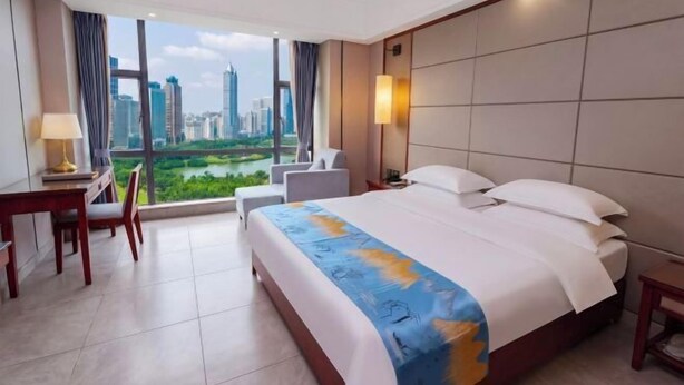 A hotel room with a large bed, a desk, a chair, a lamp, and a view of a cityscape. - Yihao Hotel (Qionghai)