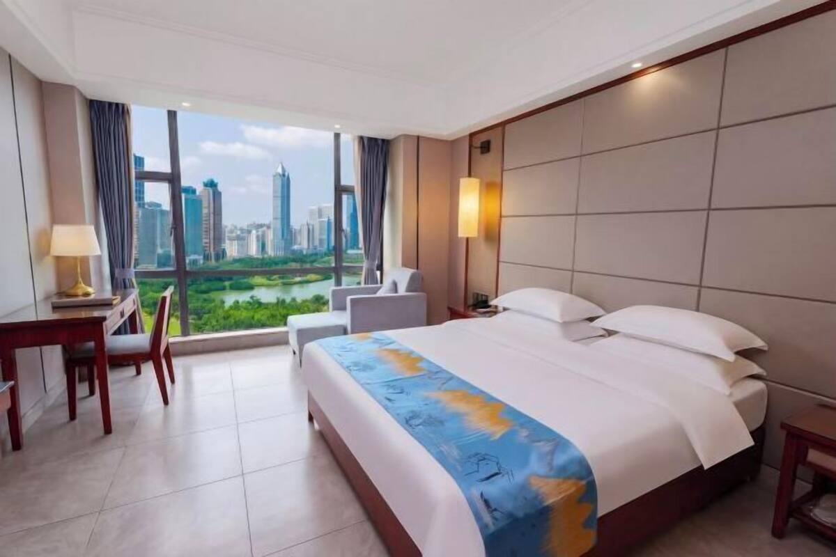 A hotel room with a large bed, a desk, a chair, a lamp, and a view of a cityscape.
