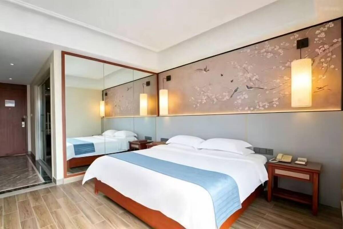 A hotel room with a large bed, a nightstand, a mirror, and a wall-mounted light fixture.