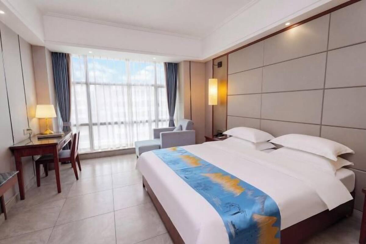 A hotel room with a large bed, a desk with a lamp, a chair, and a view of the outdoors through large windows.